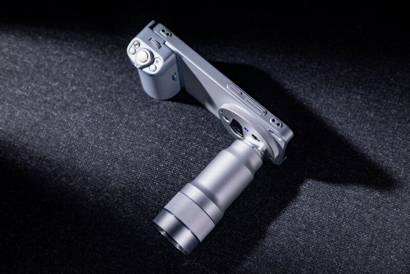 A silver compact digital camera with a large, extended zoom lens is positioned at an angle on a dark textured surface, illuminated by a focused beam of light.