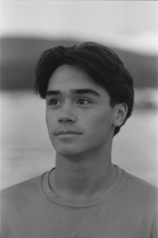 Black and white portrait of a young person with short, dark hair, looking slightly upward. The background is blurred, showing water and distant hills or mountains. The person is wearing a plain T-shirt.