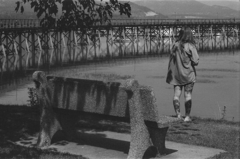 A person with long hair and tattoos on their legs stands near a riverside, facing away from the camera. A stone bench sits on the grass in the foreground, and a wooden bridge stretches across the water in the background.