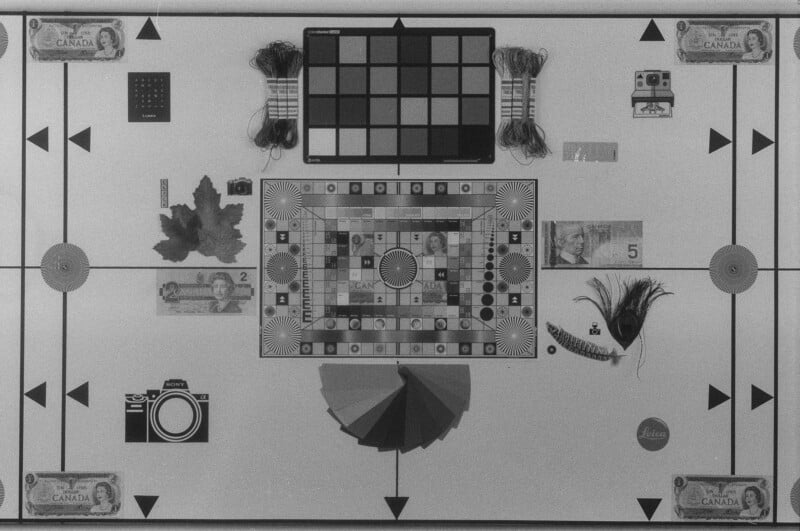 Black and white photo of a flat layout featuring currency, game pieces, leaves, feathers, cameras, thread spools, a color chart, and geometric patterns arranged symmetrically on a patterned board.