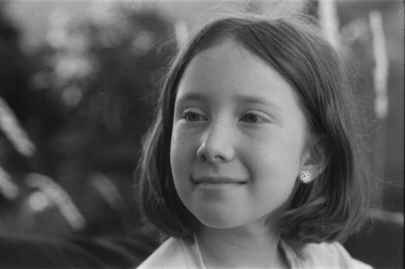 A black and white photo of a young girl with short hair, wearing a flower-shaped earring, looking slightly to the left and smiling softly. The background is blurred with natural light.