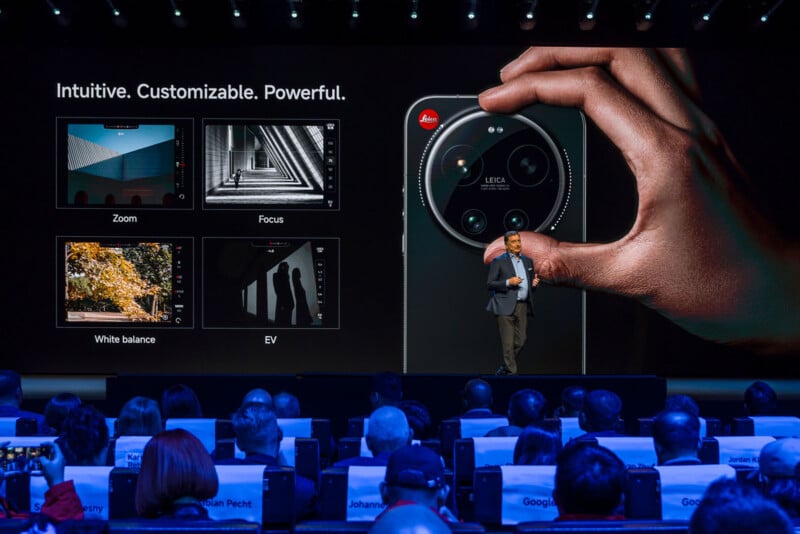 A presenter stands on stage holding a large smartphone, with a screen behind him displaying features like zoom, focus, white balance, and EV. An audience sits facing the stage in a dark auditorium.