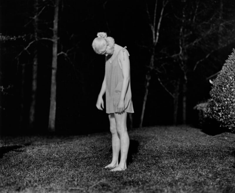 A person with light hair, wearing a sleeveless dress, stands barefoot on grass at night, head and arms hanging down, surrounded by trees and bushes in the dark background.