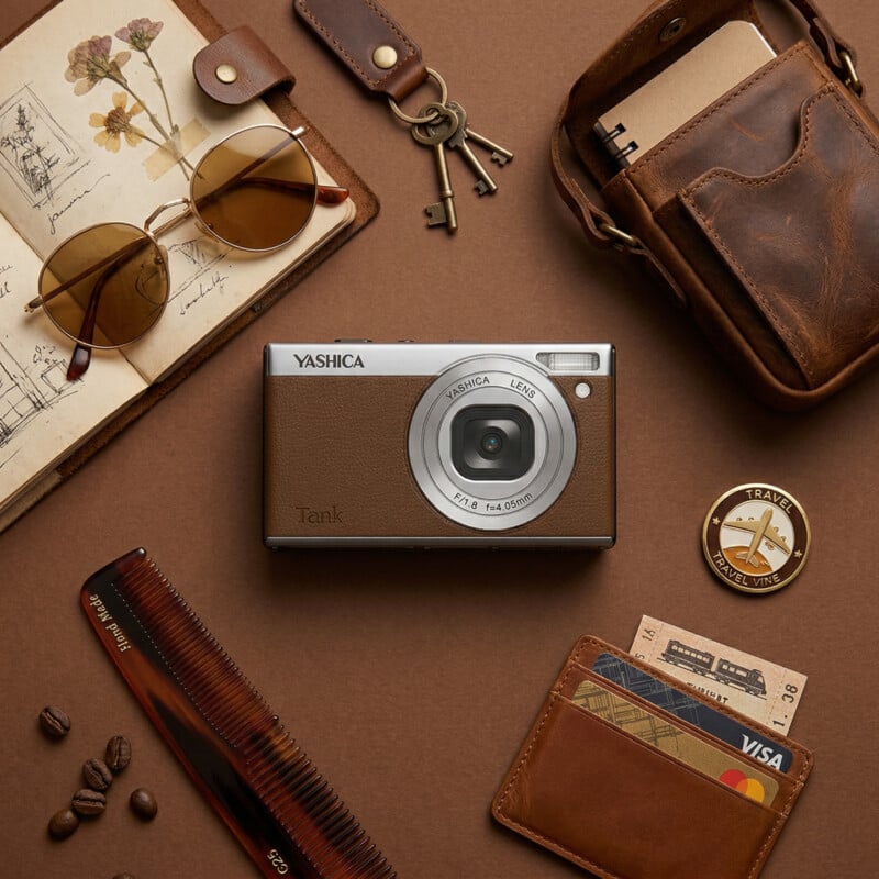 Flat lay of travel essentials on a brown surface, including a vintage camera, sunglasses, leather wallet, card holder with cards, a notebook, a compass, a comb, and coffee beans.