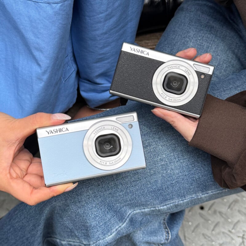 Two people sitting side by side hold Yashica digital cameras, one in light blue and the other in black, showcasing the cameras in their hands. Denim jeans and casual clothing are visible.