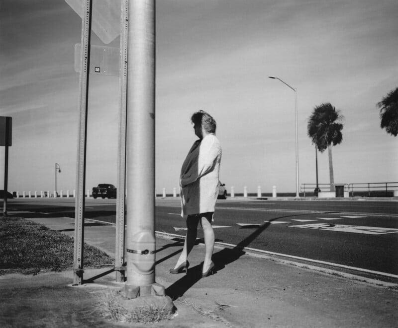 A person stands on a sidewalk near a street corner, partially shaded by a pole. The street behind is mostly empty, with a car in the distance and two palm trees along the roadside under a clear sky.