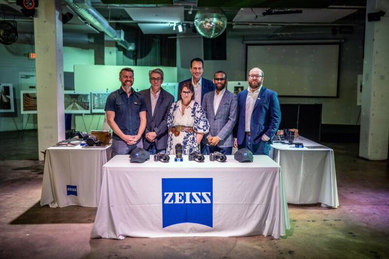 En una sala moderna de estilo industrial, seis personas se encuentran detrás de una mesa Zeiss que muestra cámaras y equipos, con una bola de discoteca encima. El grupo está sonriendo y vestido con ropa informal o elegante.