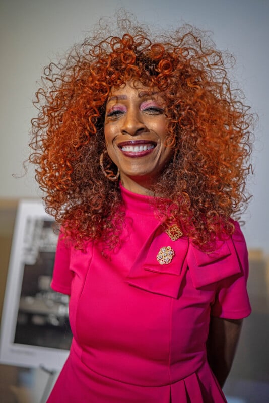 Una mujer con cabello rizado rojo vibrante y una sonrisa brillante, vestida con un vestido rosa brillante con un gran lazo y un broche de joyería, está parada en el interior con un fondo borroso.