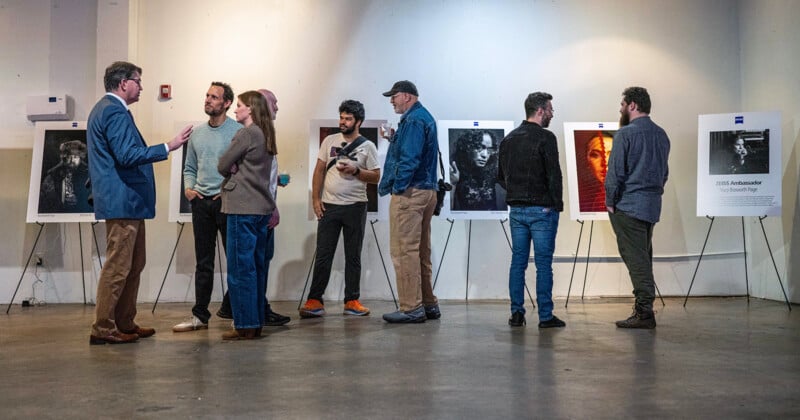 Un grupo de personas paradas y hablando en una galería de arte mirando retratos expuestos en caballetes sobre una pared blanca. El ambiente es relajado y social.