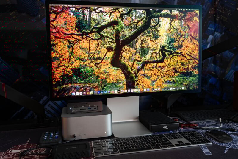 A desktop computer setup with a large monitor displaying a vibrant autumn tree, accompanied by a Mac Studio, keyboard, mouse, and various accessories on a desk.