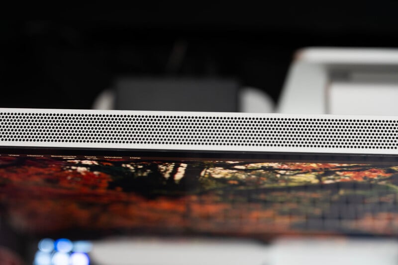 Close-up of the top edge of a computer monitor with a perforated silver speaker grille above a blurry screen showing an autumn landscape in warm tones.