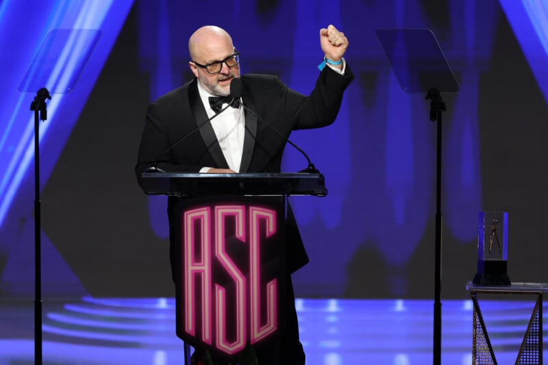 Un hombre con esmoquin se encuentra en el podio. "ASC" Frente a un escenario iluminado por luces azules y violetas, levantó su puño derecho y habló por un micrófono.