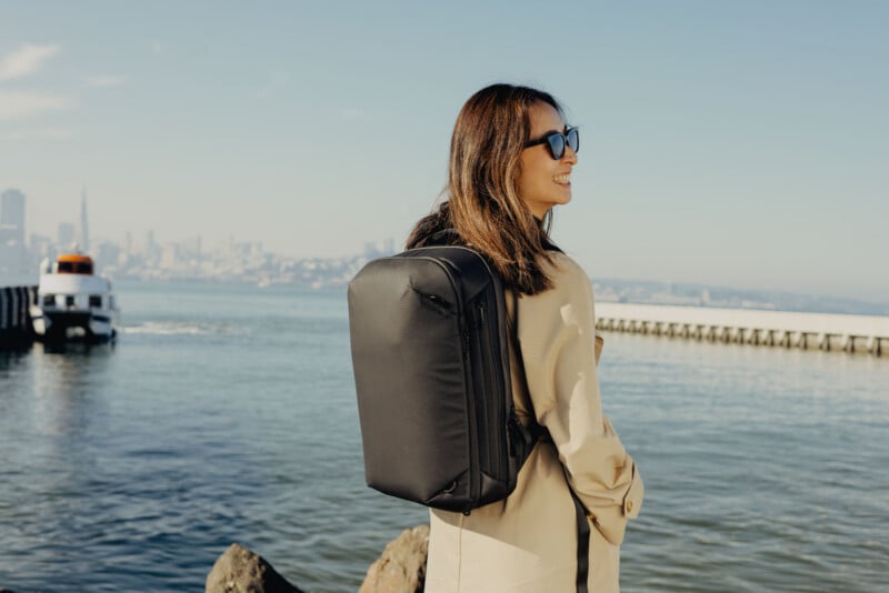 A woman wearing sunglasses and a beige coat stands by the water with a black backpack, smiling while looking toward the city skyline and a docked ferry in the background.