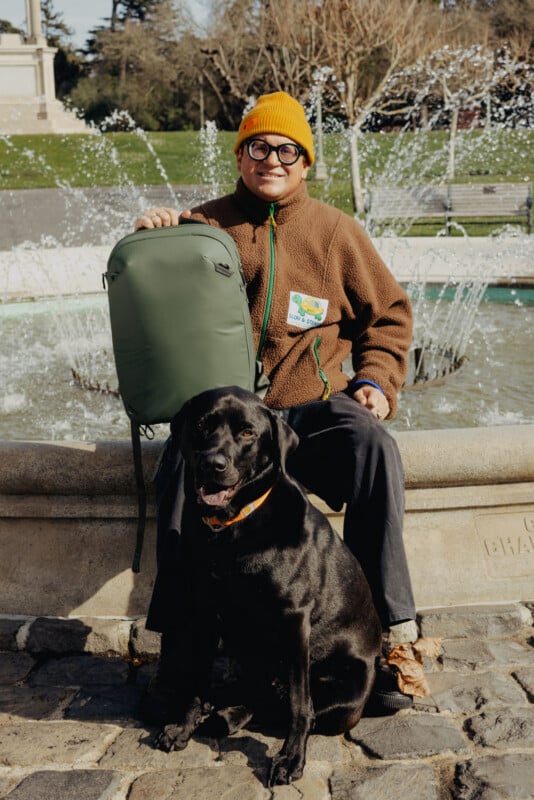 A person wearing a brown jacket, orange beanie, and glasses sits on a stone bench in front of a fountain, holding a green backpack. A black Labrador dog with an orange collar sits in front of them.
