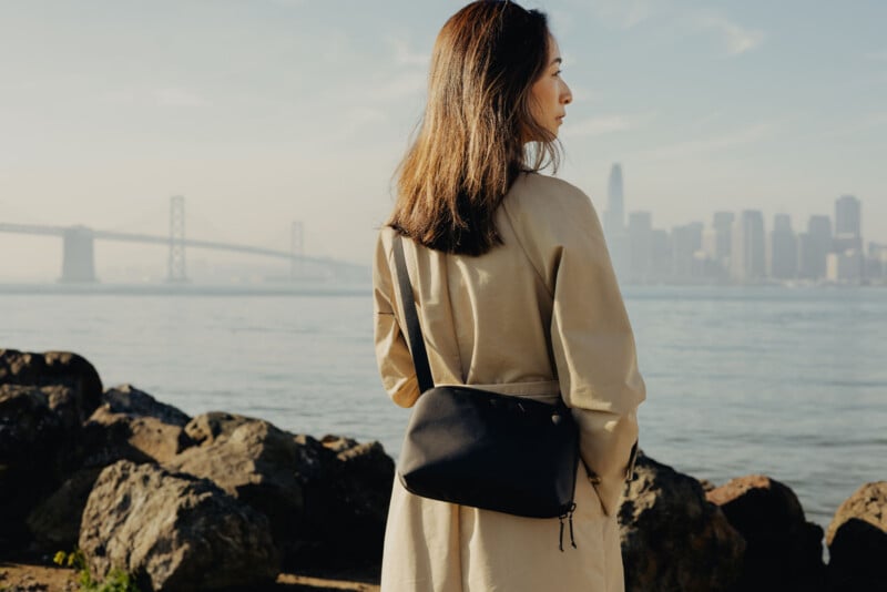A woman in a beige coat stands on rocky ground near the water, facing a city skyline and a bridge in the background on a hazy day. She has a black bag slung over her shoulder.