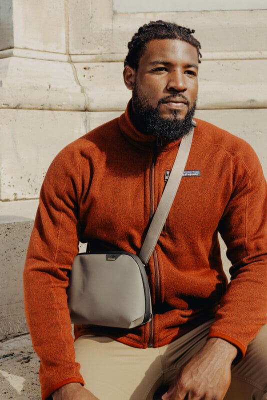 A man with a beard and short dreadlocks sits outdoors against a stone wall, wearing an orange fleece jacket and tan pants. He has a gray crossbody bag across his chest and looks off-camera with a calm expression.