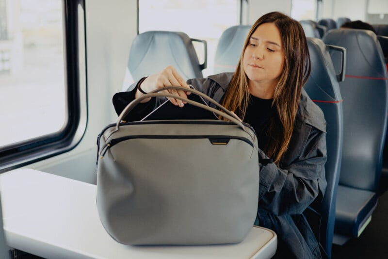 A woman sits by a window on a train, placing her hand inside a large gray bag on the table in front of her. She looks focused and wears a dark jacket.
