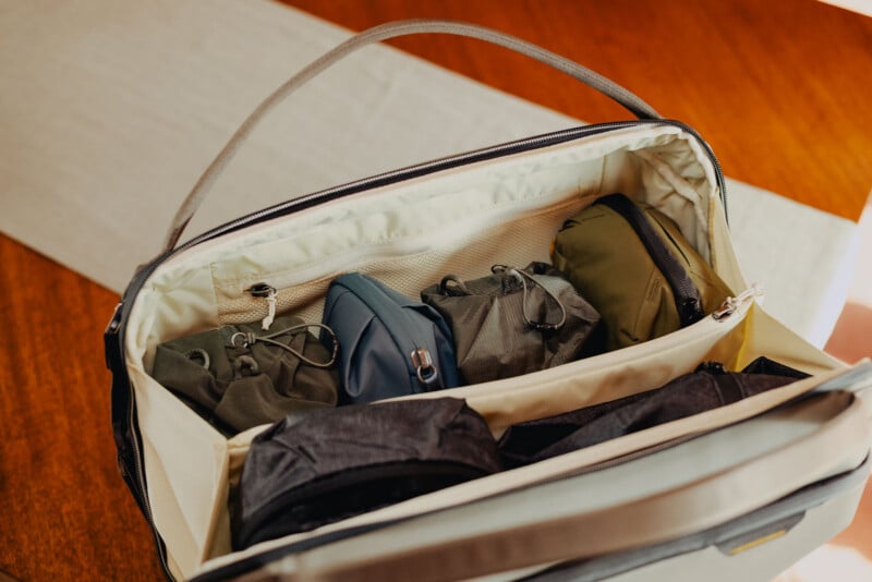 An open travel bag sits on a wooden table, neatly packed with several organized pouches and compartments holding various items. The bag has a light interior and a sturdy handle.