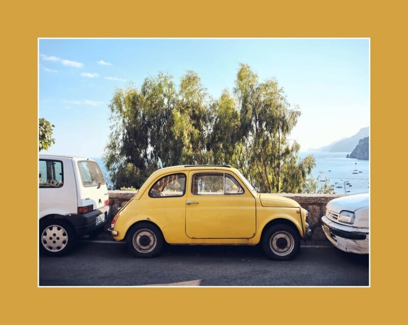 A vintage yellow car is parked between two other cars along a seaside road, with trees, blue sky, and distant mountains visible in the background. Image is framed by a wide mustard-yellow border.