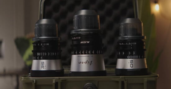 Three silver and black camera lenses are displayed upright in an olive green hard case, with blurred indoor background lighting.