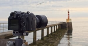 A camera on a tripod is set up on a pier, pointing toward a distant lighthouse with red and white stripes. The sky is cloudy, and a bird is flying above the water near the lighthouse.