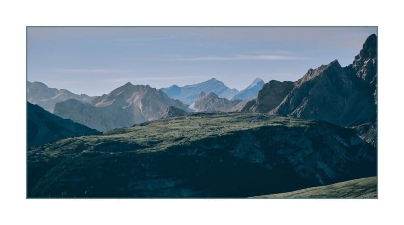 A panoramic view of rugged mountain peaks under a clear blue sky, with sunlight casting shadows across the green, sloping foreground.