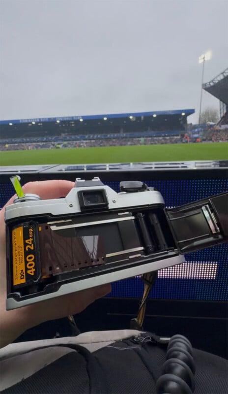 A person holds an open film camera loaded with a 400 ISO film canister at a sports stadium, with the pitch and blurred spectators visible in the background.