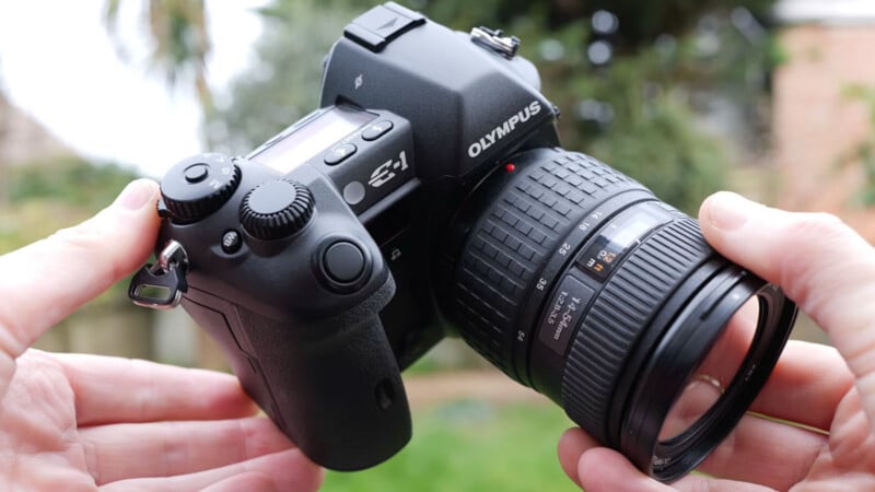 Un hombre sosteniendo una cámara digital Olympus E-1 negra con una lente grande al aire libre sobre un fondo verde y marrón borroso.