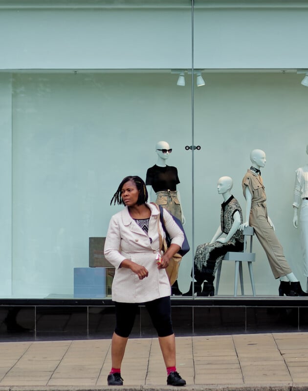 A woman stands on a sidewalk in front of a store window displaying three mannequins in fashionable outfits. She wears a light jacket, black leggings, and sneakers, and looks off to the side while holding a bag.