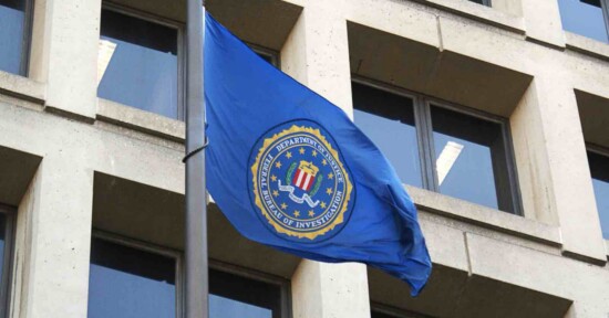 A blue flag with the FBI seal waves on a flagpole in front of a beige office building with large windows.