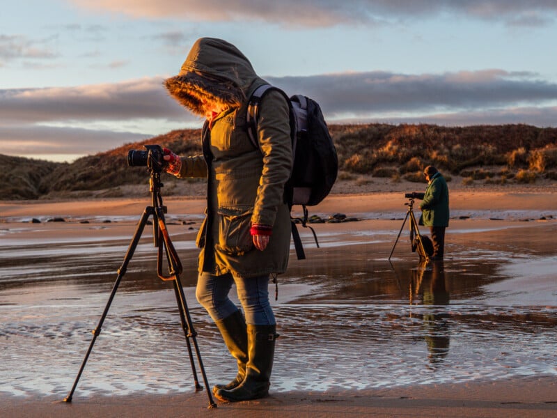 Two people with tripods and cameras stand on a wet, sandy beach at sunset. The person in the foreground wears a hooded parka and boots, while the person behind also photographs the scene. Low hills and clouds are in the background.