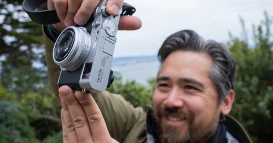 A smiling man holds up a silver Fujifilm camera outdoors, with greenery and water blurred in the background.