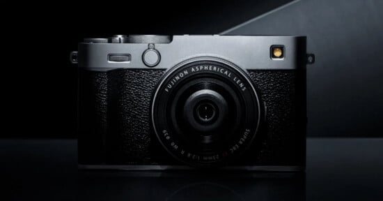 A Fujifilm digital camera with a black textured body and prominent lens is shown on a dark, reflective surface against a dark background.
