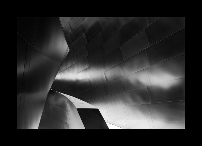Black and white photo of curved, reflective metal panels forming an abstract architectural structure with soft light and shadows accentuating the flowing shapes.