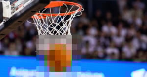 A basketball is about to go through a hoop with a net in an indoor arena; the ball is pixelated and the background shows a blurred crowd.
