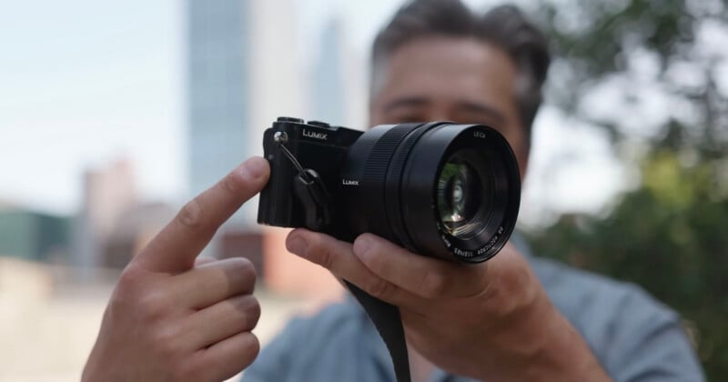 A person holds a Lumix camera with one hand and points at it with the other, with a blurred urban background and greenery behind them.