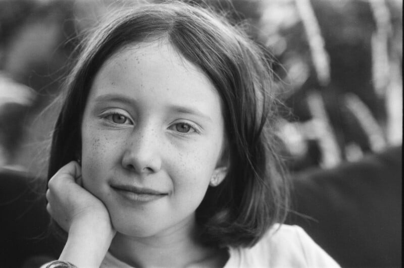 Black and white photo of a young girl with shoulder-length hair and freckles, resting her chin on her hand, looking at the camera with a gentle smile. Blurred background with natural light.