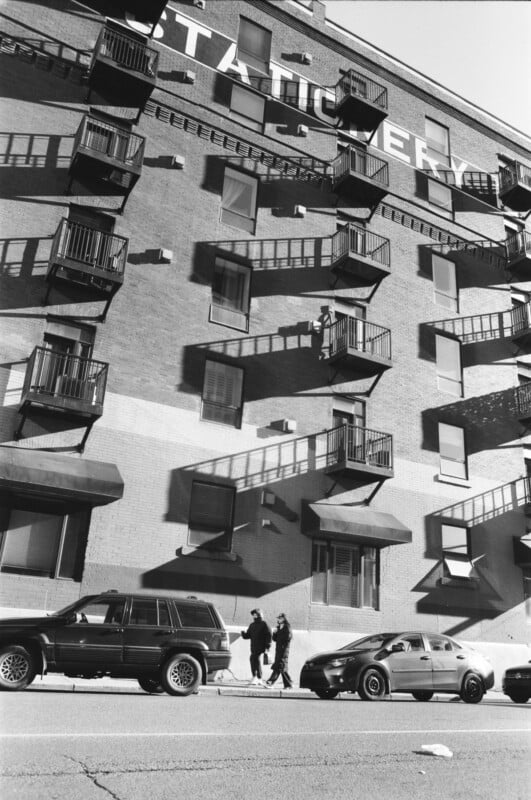 Black and white photo of a brick apartment building with balconies casting shadows. Two cars are parked in front, and two people walk together on the sidewalk, crossing in front of the vehicles.