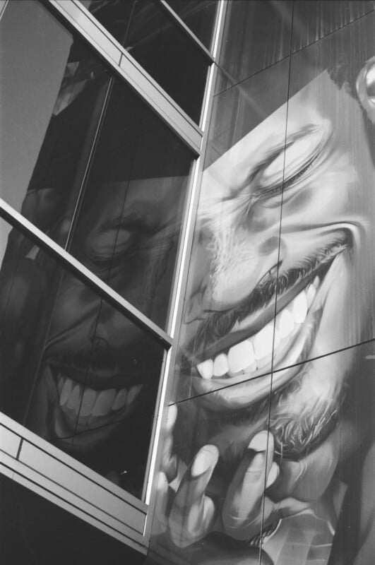 Black and white photo of a building facade with a large mural of a person’s smiling face, distorted by the angles of the window panes, emphasizing expressive eyes and a wide grin.