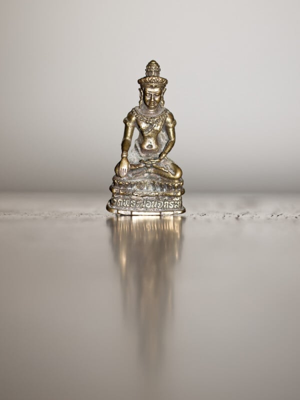 A small metallic Buddha statue sits on a reflective surface, creating a subtle reflection. The statue features intricate detailing and Thai script at the base, with a plain, blurred background.