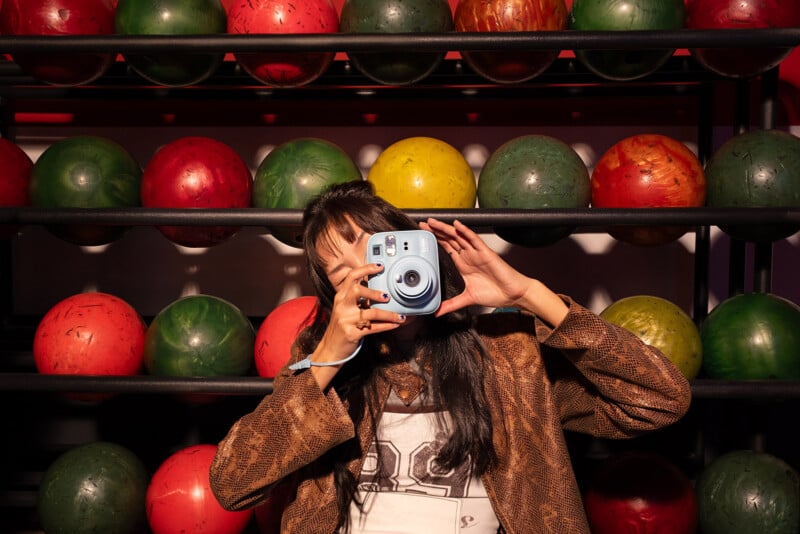 Un hombre se sienta frente a una colorida bola de bolos, sosteniendo una cámara instantánea azul frente a su cara como si estuviera tomando una fotografía. El fondo está lleno de bolas de bolos rojas, verdes y amarillas dispuestas sobre una rejilla.