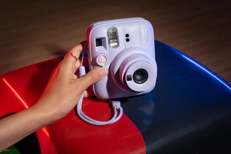 Sosteniendo una cámara instantánea Fujifilm Instax Mini 11 de color violeta claro (viene con correa para la muñeca) en una mano, apoyada sobre una superficie colorida y brillante con secciones rojas y azules.