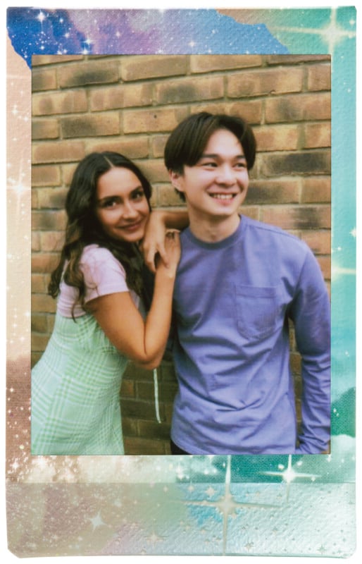 Dos personas sonriendo para una foto frente a una pared de ladrillos. La persona de la izquierda lleva un vestido a cuadros verde con una camisa blanca debajo, apoyada contra la persona a su lado que lleva una camisa de manga larga azul claro. La foto tiene un borde colorido y brillante.