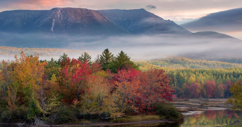 Colorful autumn trees with red, orange, and yellow leaves stand by a calm lake, with mist rising in front of forested hills and a tall, rugged mountain under a partly cloudy sky.