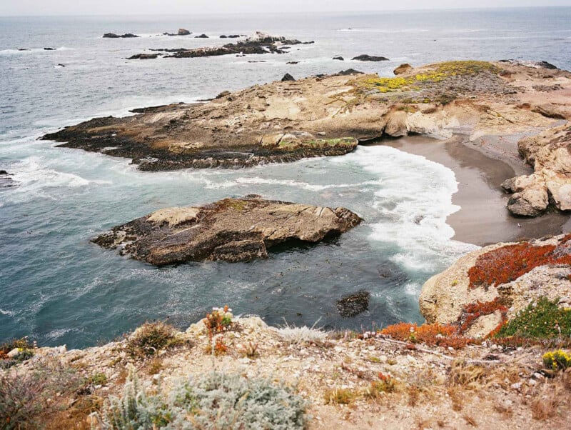 Costa rocosa con pequeñas calas de arena, olas que baten suavemente y una mancha de vegetación verde y roja en primer plano. El cielo estaba cubierto de nubes oscuras y en la costa se veían varias islas rocosas.