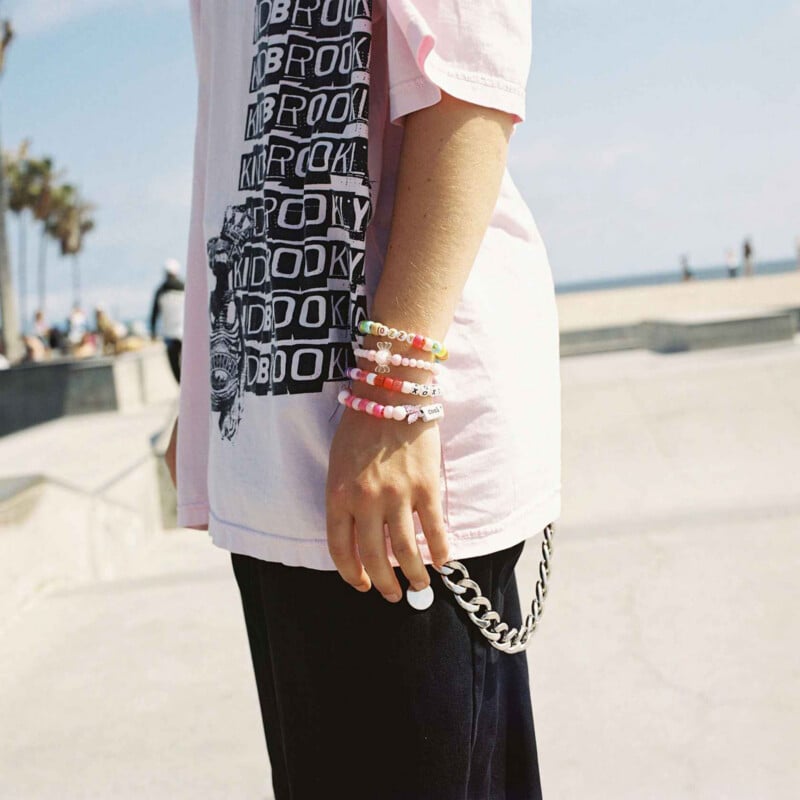Un hombre vestido con una camiseta rosa, pantalones negros, una cadena de plata y una pulsera de cuentas de colores se encuentra al aire libre cerca de la pista de patinaje de la playa con palmeras y un cielo azul de fondo.