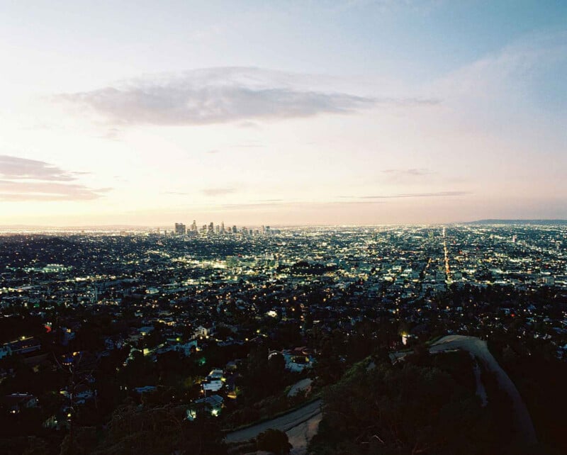 El paisaje urbano al anochecer es vasto, con innumerables luces iluminando los edificios y las calles. A lo lejos se ve el centro de la ciudad con sus altos edificios, con algunas nubes dispersas bajo el cielo.