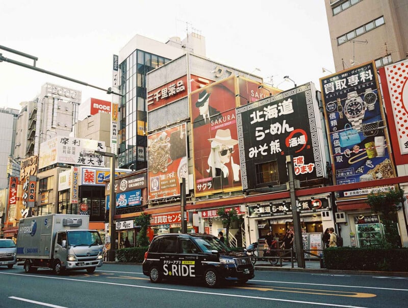 Coloridas calles de la ciudad japonesa repletas de tiendas, grandes carteles publicitarios y carteles japoneses. Por la calle circulaban un taxi negro y una furgoneta de reparto. Las calles están llenas de edificios altos y escaparates concurridos.