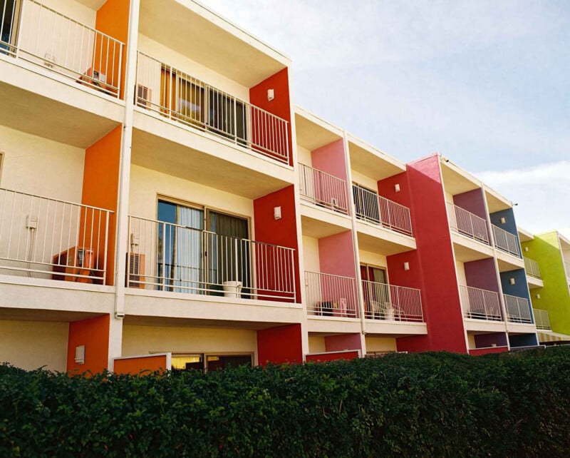 Un moderno edificio de apartamentos de tres pisos con balcones privados en cada piso. Las paredes exteriores están pintadas en colores brillantes como naranja, rojo, rosa y verde. Setos verdes bordean la base del edificio.