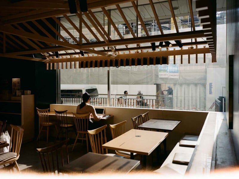 Un hombre estaba sentado solo en una mesa de un café tranquilo y soleado, con sillas de madera y grandes ventanales, mirando a la gente y los vehículos que pasaban por la calle.